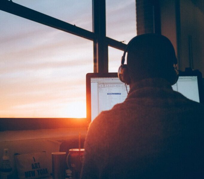 Eine Person sitzt vor einem Computerbildschirm, der in einem warmen, goldenen Licht erleuchtet wird. Durch ein großes Fenster im Hintergrund ist ein ruhiger Sonnenuntergang zu sehen, der den Raum in sanfte Farben taucht. Die Person trägt Kopfhörer und wirkt konzentriert, während der Abendhimmel allmählich dunkler wird.